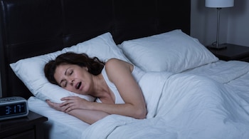 Una mujer con cabello oscuro duerme en una cama con sábanas blancas con la boca abierta junto a un reloj digital que marca las tres y diecisiete.