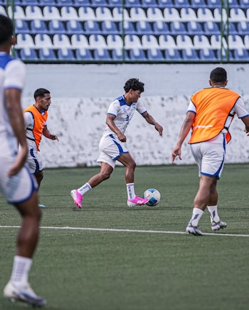 Un futbolista con uniforme blanco conduce el balón sobre el césped mientras otros jugadores en chalecos naranjas lo observan en un estadio