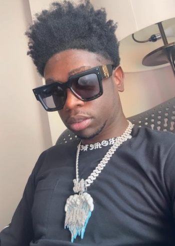 Un hombre joven con gafas de sol oscuras, camiseta negra y una cadena plateada con un colgante con forma de nube