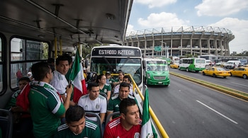 Vista desde el interior de un autobús con aficionados mexicanos. Otro autobús con destino al Estadio Azteca se ve al frente, con el estadio al fondo y tráfico.