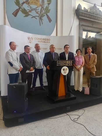 Siete personas, seis hombres y una mujer, en un escenario con podio y altavoces. Fondo blanco con logotipo 'Congreso de la República'