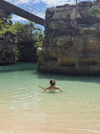Una mujer de espaldas en el agua clara de una laguna, rodeada de paredes de piedra y con una pasarela de madera en la parte superior