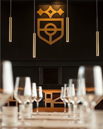 Copas de vino en primer plano desenfocado con un logo dorado en forma de escudo y luces colgantes en una pared oscura del Restaurante Lautaro Martinez