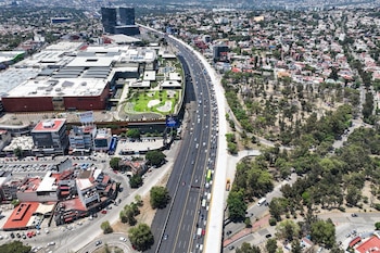 Vista aérea de una autopista llena de vehículos, un centro comercial grande con techos verdes y un parque arbolado, con edificios residenciales al fondo