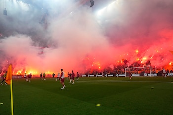 Vista de un campo de fútbol con jugadores entrenando; las gradas al fondo están llenas de aficionados con bengalas rojas y humo intenso. Hay un banderín naranja en primer plano