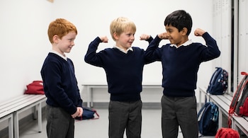 Tres niños varones de uniforme escolar posan flexionando los brazos en un vestuario, con bancos y mochilas al fondo.