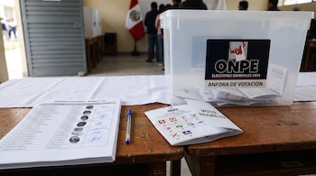 Un padrón electoral con huellas, un bolígrafo azul, una cédula de votación y un ánfora transparente sobre una mesa de madera.