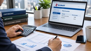 Persona en un escritorio frente a una computadora portátil con la página web de SUNAT, documentos contables impresos y una calculadora