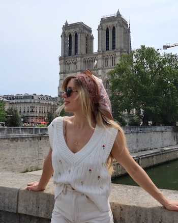 Mujer rubia con pañuelo y gafas de sol recostada en un puente con la Catedral de Notre Dame, edificios y un río al fondo