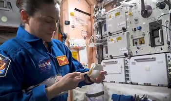 Una mujer con uniforme azul de la NASA sostiene un recipiente blanco y una boquilla plástica dentro de un módulo espacial con paneles y cables