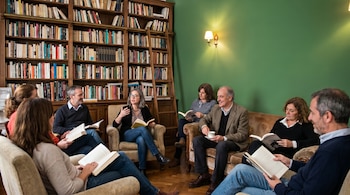 Ocho personas adultas sentadas en un salón, rodeadas de libros en una estantería grande. La pared es verde inglés con una lámpara de aplique encendida.