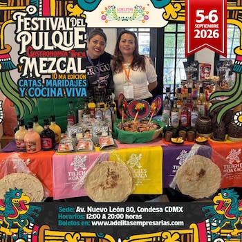 Dos mujeres posan detrás de una mesa con botellas de mezcal, tortillas artesanales y productos tradicionales debajo del anuncio de un festival