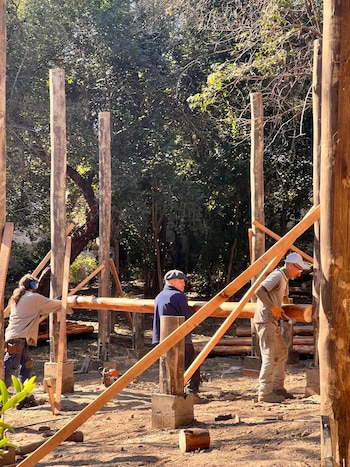 Tres personas construyen una estructura de madera. Se observan postes, vigas y herramientas en un terreno de tierra con árboles al fondo