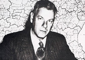 Retrato en blanco y negro de Hendrik Frensch Verwoerd, un hombre de mediana edad con traje de rayas y corbata a cuadros, delante de un mapa