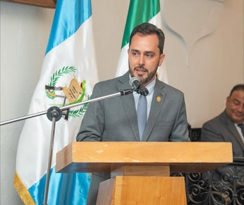 Un hombre con traje gris y corbata habla ante un micrófono en un podio de madera frente a la bandera de Guatemala