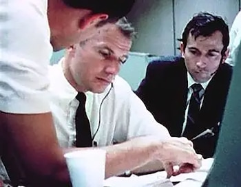 Dos hombres concentrados en un centro de control. Uno con camisa blanca y corbata, otro con traje oscuro, ambos con auriculares, revisan documentos