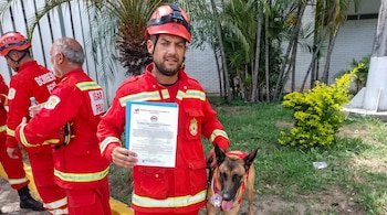 Bombero peruano en uniforme rojo y casco sujeta un documento, junto a un perro pastor belga malinois con chaleco y medallas. Otros bomberos, pared y palmeras.