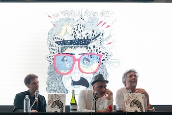 Tres hombres en una mesa, con Fito Páez a la derecha, frente a una pantalla mostrando una ilustración de una cara con gafas y edificios. Libros y botellas en la mesa