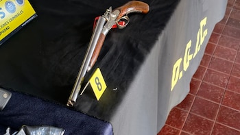 Un arma de fuego de dos cañones reposa sobre una mesa cubierta con una manta negra junto a un marcador amarillo con el número ocho