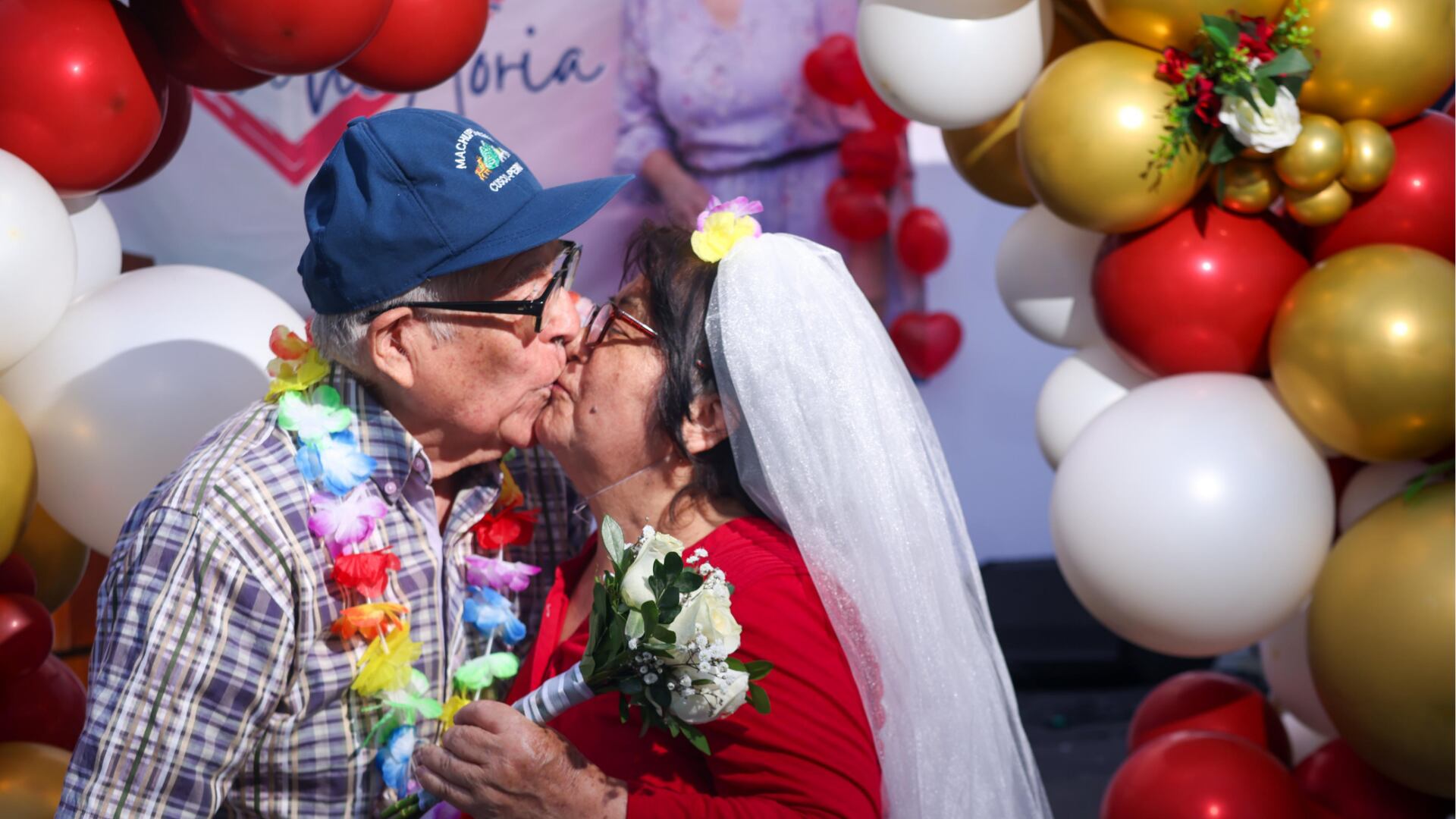Una pareja de adultos mayores comparte un tierno beso mientras renuevan sus votos matrimoniales en una conmovedora ceremonia celebrada durante el Día de San Valentín, rodeados de globos festivos.
