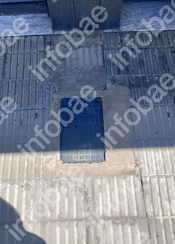 Tapa de servicio rectangular oscura con la palabra "AGUA" en relieve, rodeada de baldosas de hormigón gris con textura y grietas, en la entrada de un edificio