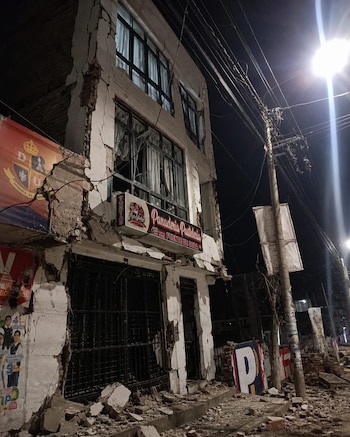 Edificio de tres pisos con daños en la fachada, un letrero de 'Panadería Pastelería', escombros en la acera, poste con cables y farola de noche