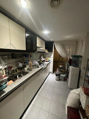 Cocina estrecha con gabinetes superiores blancos y negros, encimera de granito con fregadero y platos. Se ven una estufa, suelo de baldosas grises y un dispensador de agua