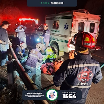Bombero de espalda con casco rojo observa a rescatistas y paramédicos asistiendo a una persona junto a una ambulancia S.P. Jocopilas en carretera oscura