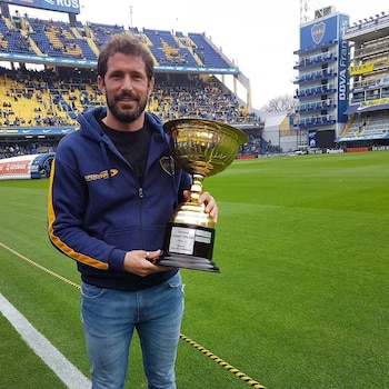 Un hombre con barba y sudadera azul sostiene un trofeo dorado grande en un campo de fútbol. Detrás se ven gradas azules y amarillas con público y publicidad