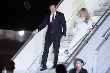 Un hombre con traje y corbata roja y una mujer vestida de blanco bajan por la escalinata de un avión durante la noche