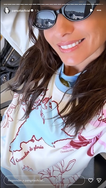 Pampita sonríe con cabello castaño suelto y gafas de sol que reflejan un avión en la pista. Viste un suéter claro con un diseño