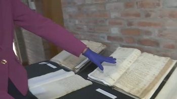 Una persona con saco morado y guante azul señala un libro antiguo abierto sobre una mesa con otros documentos expuestos