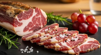 Un trozo de solomillo de cerdo curado con varias rebanadas finas a su lado, espolvoreadas con sal marina y pimienta. Se ven ramitas de romero y uvas rojas en una tabla oscura.