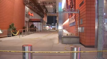Una calle exterior de noche acordonada con cinta amarilla de "escena del crimen" frente a edificios con fachadas naranjas y letreros iluminados