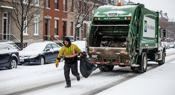 Un recolector de basura con chaqueta amarilla de alta visibilidad corre en una calle nevada con una bolsa negra, detrás de un camión de basura verde en Montreal.
