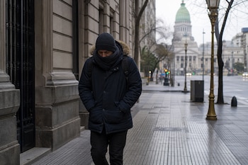 Un hombre con abrigo con capucha, gorro y bufanda camina por la acera cerca de un edificio con cúpula.