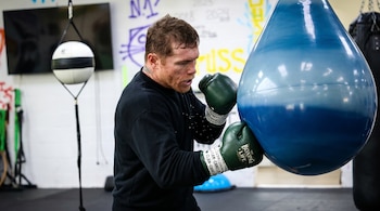 Saúl Canelo Álvarez con guantes verdes golpea un saco de boxeo azul dentro de un gimnasio.