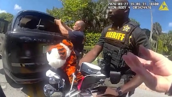 Un oficial de sheriff en uniforme con chaleco antibalas amarillo y negro se para junto a un Range Rover negro con la ventana trasera rota. Palos de golf en primer plano