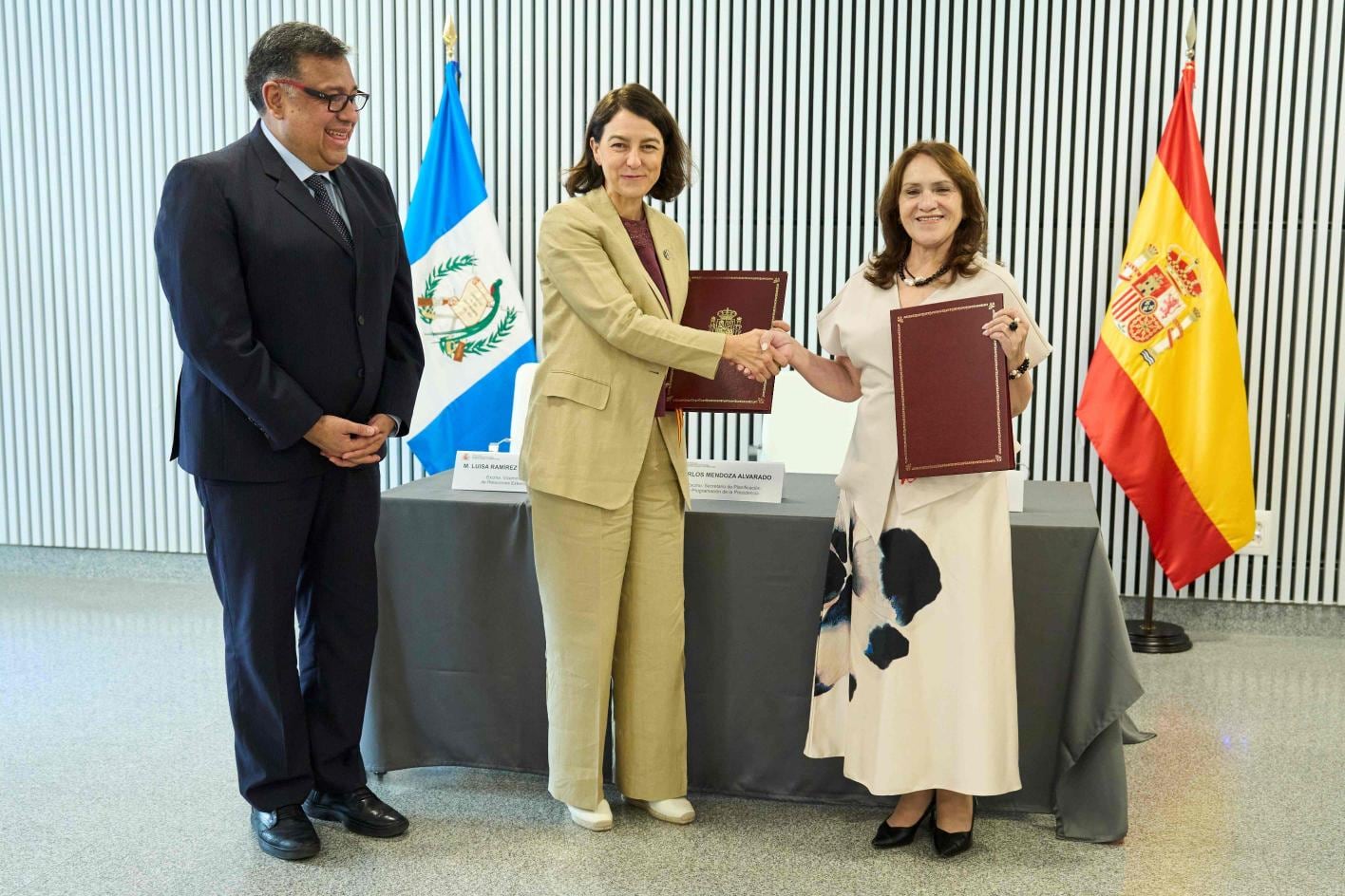 Una funcionaria de Guatemala y otra de España se saludan tras firmar un acuerdo bilateral, con un observador presente. (Ministerio de Relaciones Exteriores de Guatemala)