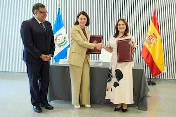 Tres personas, dos mujeres y un hombre, en un evento. Las mujeres se dan la mano y sostienen carpetas. Banderas de Guatemala y España detrás