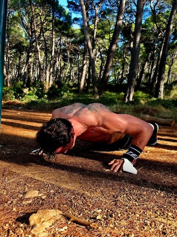 Un hombre con el torso desnudo y tatuajes en la espalda hace flexiones de brazos sobre el suelo en un bosque de pinos
