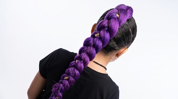 Vista posterior de una niña con camiseta negra y una trenza gruesa de color morado con anillos dorados sobre un fondo blanco.