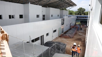 Edificios modulares blancos en construcción, con un techo a la vista. Terreno de tierra, dos trabajadores con cascos y chalecos. Árboles verdes al fondo
