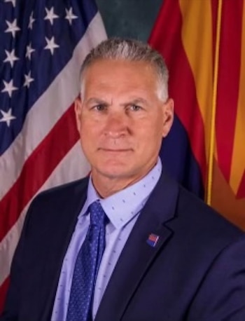 Retrato de Frank Strada con traje azul, camisa clara y corbata, delante de la bandera de Estados Unidos y la bandera de Arizona