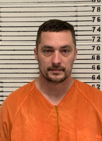 Fotografía policial de un hombre de piel clara con barba, cabello oscuro corto y vistiendo un uniforme naranja de prisión, parado frente a un medidor de altura
