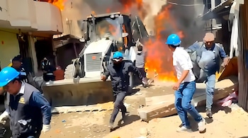Hombres con cascos azules corren en una calle frente a una maquinaria de construcción rodeada por fuego y humo.