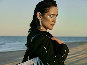 Julieta Venegas, con maquillaje brillante y traje oscuro con perlas, lleva un acordeón Gabbanelli negro en una playa al atardecer, con el mar