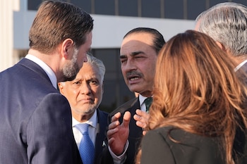 Primer plano de un hombre con bigote y traje oscuro gesticulando mientras habla. Un hombre de espalda parcial y un hombre mayor con barba gris lo escuchan. Una mujer de espalda también se ve