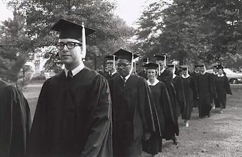 Fotografía en blanco y negro de estudiantes con togas y birretes caminando en fila por un terreno al aire libre con árboles