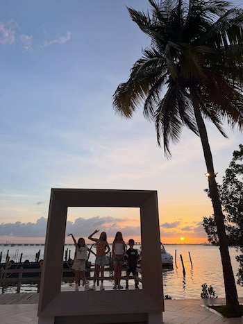 Cuatro niños posan dentro de un marco decorativo al atardecer, con un muelle, botes, palmeras y el sol poniéndose sobre el agua de fondo